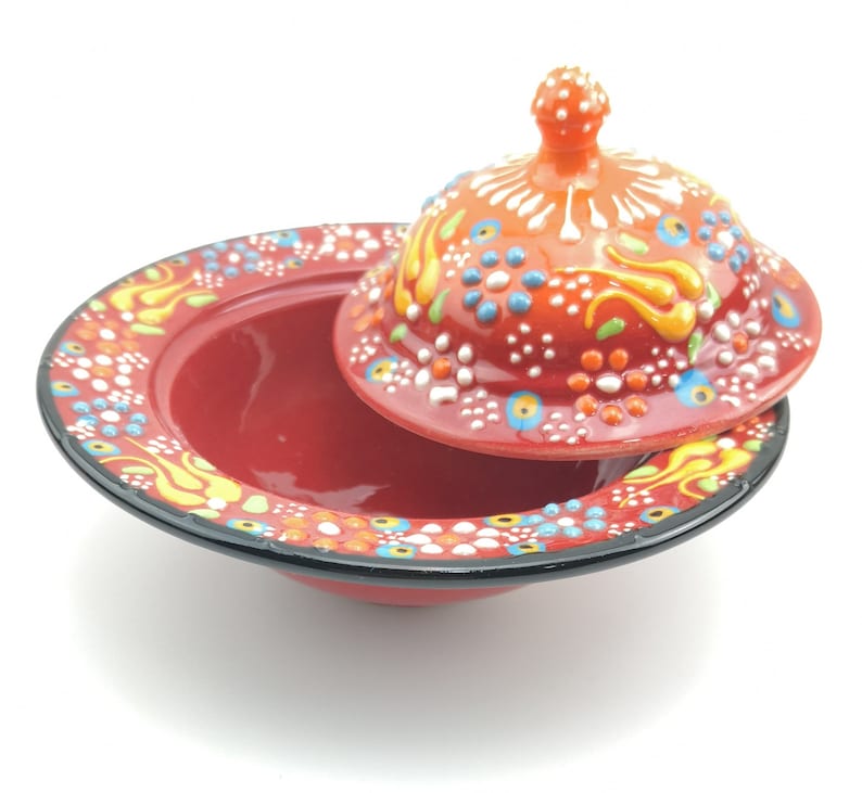 May include: A red ceramic dish with a lid, both decorated with colourful floral patterns and white dots. The lid has a small knob on top.