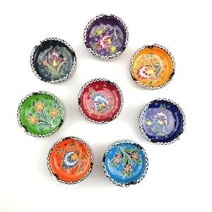 Può includere: Otto posacenere in ceramica colorati con motivi floreali. Ogni posacenere ha un colore e un motivo diverso, tra cui verde, blu, rosso, arancione e viola.