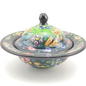 May include: A green ceramic bowl with a lid, both decorated with colourful floral patterns. The bowl has a wide, flat base and a rounded top. The lid has a knob on top.