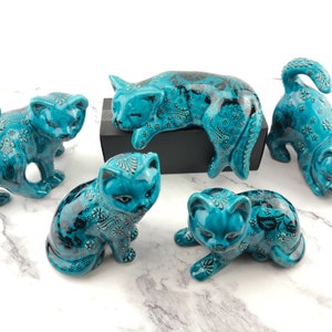 May include: Five turquoise ceramic cat figurines with black floral designs. The cats are in various poses, including sleeping, sitting, and standing.