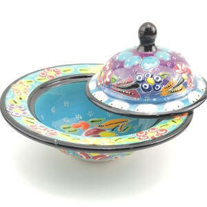 May include: A blue and white ceramic bowl with a lid, both decorated with colourful floral patterns. The bowl has a black rim and the lid has a black knob.