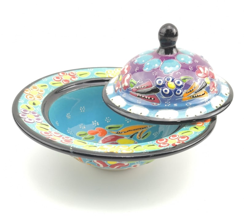 May include: A blue and white ceramic bowl with a lid, both decorated with colourful floral patterns. The bowl has a black rim and the lid has a black knob.