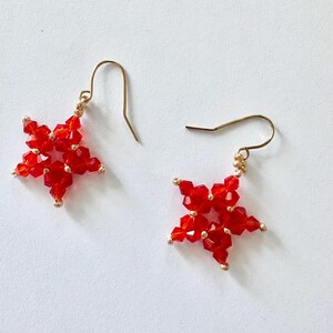 May include: A pair of star-shaped earrings. Each earring features a star design crafted from small, faceted red beads. The beads are strung together and attached to gold-colored ear wires. The earrings are displayed against a white background.