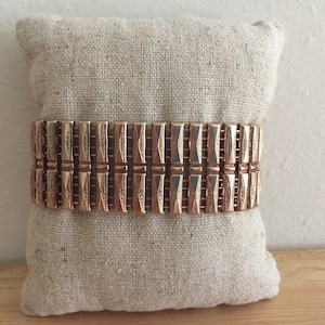 May include: A gold-toned metal bracelet with a geometric design. The bracelet is made of multiple linked pieces, each with a textured surface.