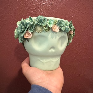 Puede incluir: Un cráneo de cerámica verde claro con una corona de flores de tela verde y rosa.