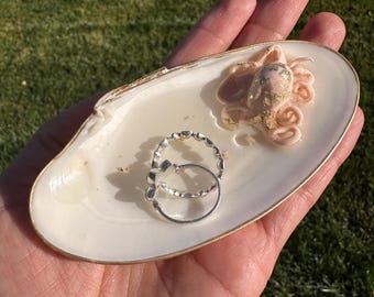 Puede incluir: Un joyero de concha blanca con borde dorado, que contiene dos anillos de plata y una pequeña figura de pulpo rosa con detalles dorados. El plato se sostiene en una mano.