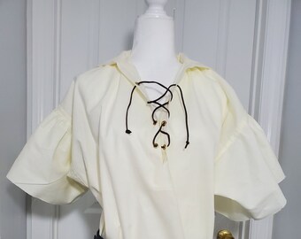 May include: A cream-colored, short-sleeved shirt with a lace-up neckline and a hood. The shirt is worn with a black leather belt with a gold buckle.