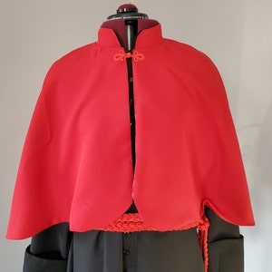 May include: A red cape with a mandarin collar and a decorative knot closure. The cape is made of a smooth fabric and has a rounded hem.
