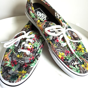Peut inclure: Une paire de baskets Vans avec un motif vibrant et coloré représentant des zèbres, des girafes et des éléments floraux. Les chaussures ont des lacets blancs, une semelle blanche et le logo Vans sur la languette.