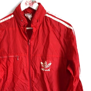 Op de afbeelding: Rode vintage trainingsjack met witte strepen op de mouwen en schouders. Voorzien van een volledige rits, twee voorzakken en het Adidas-logo op de borst. De jas hangt aan een witte hanger.