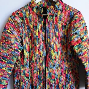 Peut inclure: Veste matelassée aux couleurs vives, ornée d'un motif multicolore. Elle est dotée d'une fermeture éclair noire sur toute la longueur, de deux poches avant et d'un col montant. Le motif est un mélange de rouge, jaune, bleu et vert.