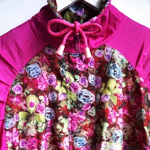 May include: A vibrant pink jacket with a floral pattern. The jacket features a high collar, a drawstring closure, and a colorful floral print with pink, purple, and blue roses. The jacket is a stylish outerwear piece.