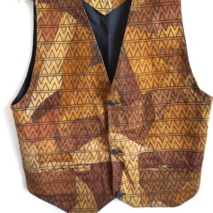 May include: A vintage brown and tan vest with a geometric pattern. The vest has a black back and lining, with three buttons down the front. The vest is hanging on a white hanger.