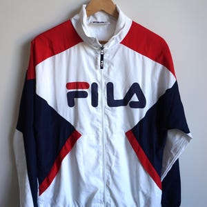 Könnte beinhalten: Eine weiße, rote und marineblaue Fila-Trainingsjacke mit durchgehendem Reißverschluss. Auf der Brust befindet sich das marineblaue Fila-Logo. Die Jacke hängt an einem Holzkleiderbügel.