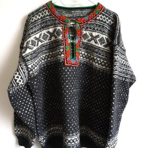Puede incluir: Un suéter de punto de manga larga en color gris oscuro y blanco con un complejo patrón geométrico. El cuello presenta un borde bordado decorativo en rojo, verde y azul con dos cierres de metal plateado en la parte central delantera.
