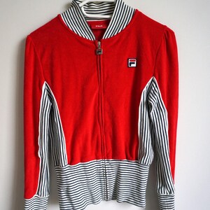 May include: A red zip-up jacket with white and black striped accents. The jacket features a white collar with black stripes, a small logo on the chest, and striped cuffs. The jacket is hanging on a white hanger.