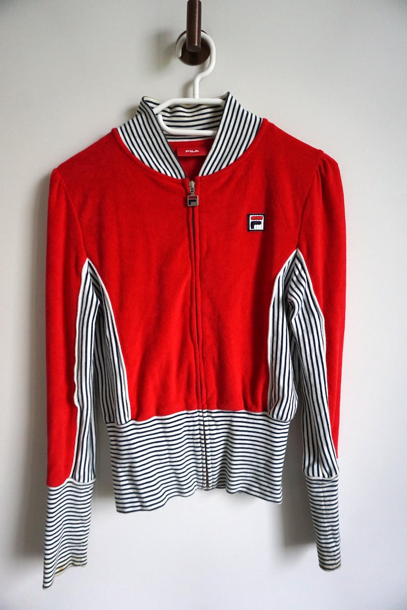 May include: A red zip-up jacket with white and black striped accents. The jacket features a white collar with black stripes, a small logo on the chest, and striped cuffs. The jacket is hanging on a white hanger.