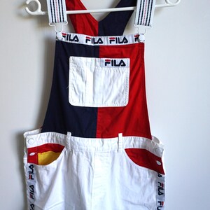 Puede incluir: Pantalones cortos con peto Fila vintage en rojo, blanco y azul marino. El peto tiene un bolsillo blanco y el logotipo de Fila. Las correas y los lados de los pantalones cortos tienen el logotipo de Fila impreso. Los pantalones cortos cuelgan de una percha blanca.