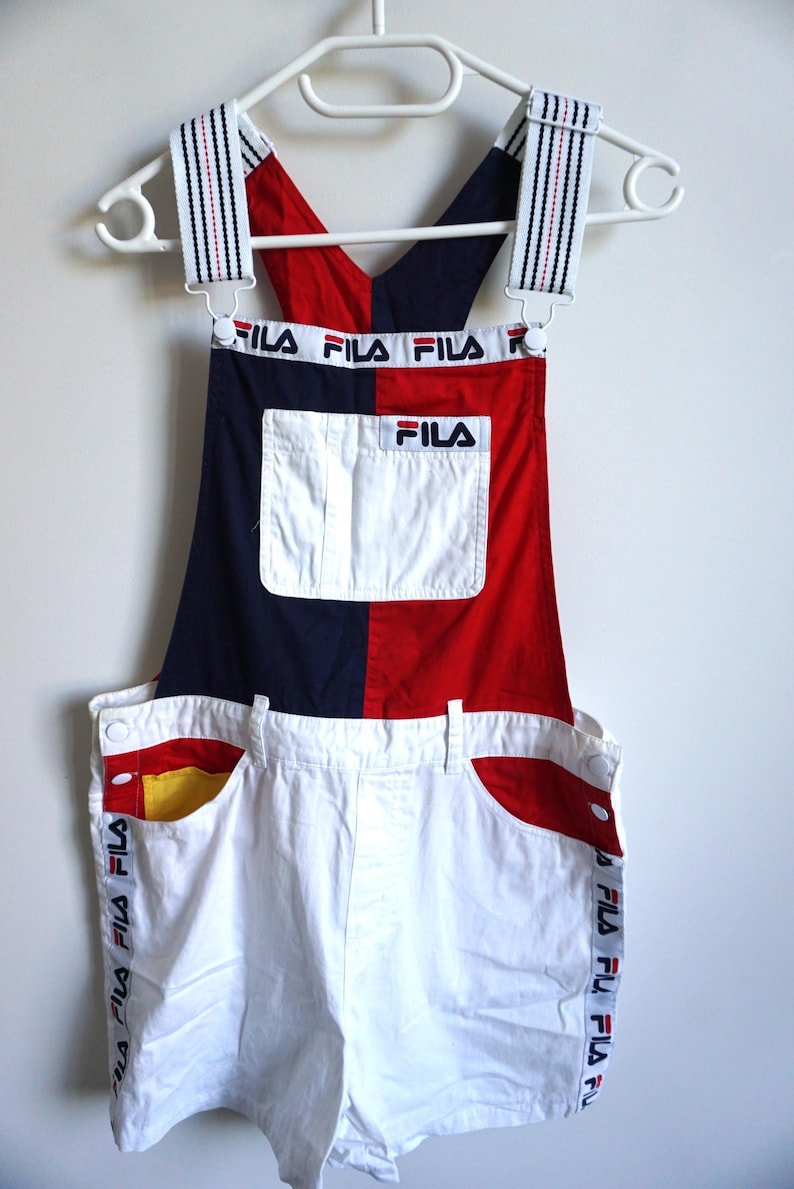 Puede incluir: Pantalones cortos con peto Fila vintage en rojo, blanco y azul marino. El peto tiene un bolsillo blanco y el logotipo de Fila. Las correas y los lados de los pantalones cortos tienen el logotipo de Fila impreso. Los pantalones cortos cuelgan de una percha blanca.