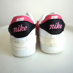 Puede incluir: Par de zapatillas Nike blancas con detalles rosas y negros. El logotipo de Nike est&aacute; bordado en rosa en el tal&oacute;n. Las zapatillas tienen un dise&ntilde;o cl&aacute;sico con suela blanca.