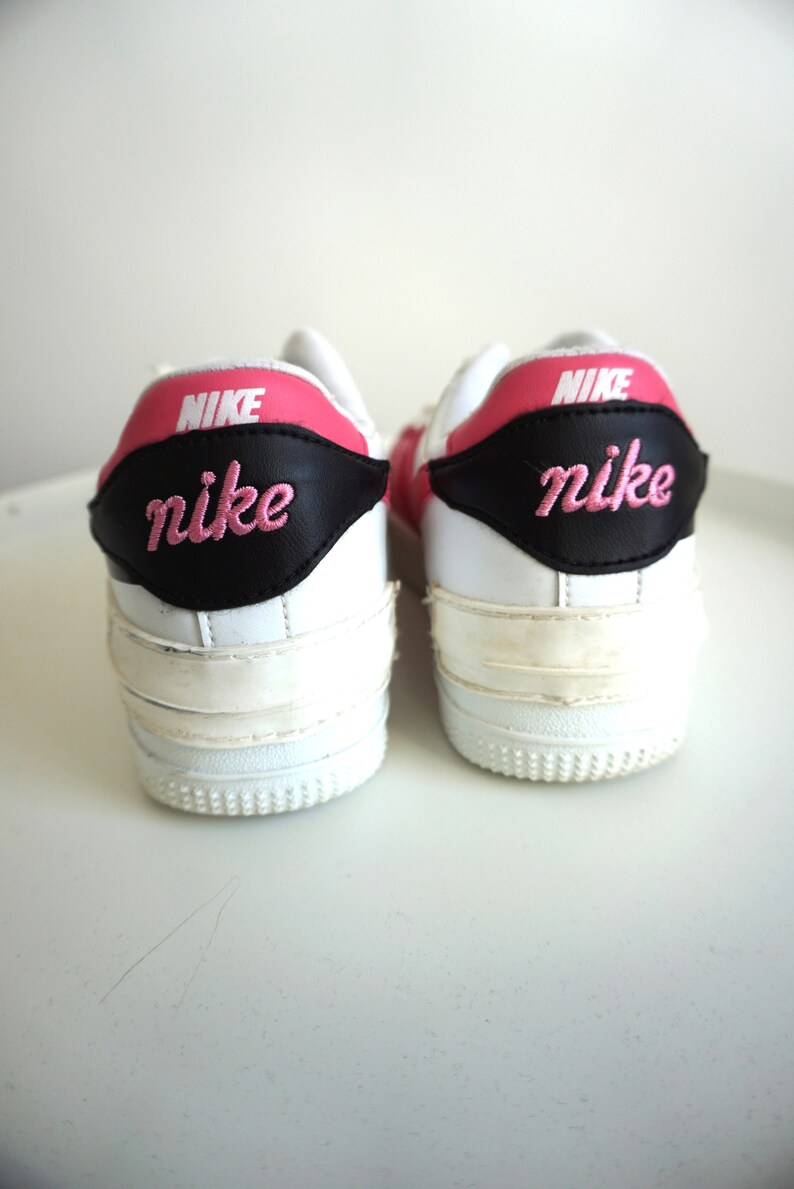 Puede incluir: Par de zapatillas Nike blancas con detalles rosas y negros. El logotipo de Nike est&aacute; bordado en rosa en el tal&oacute;n. Las zapatillas tienen un dise&ntilde;o cl&aacute;sico con suela blanca.