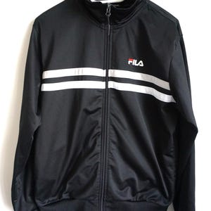 May include: A black track jacket with a high collar and a full-length front zipper. The garment features two horizontal white stripes across the chest and the Fila logo in white and red on the upper left side. It has long sleeves with ribbed cuffs.