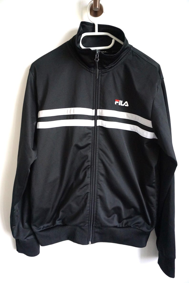 May include: A black track jacket with a high collar and a full-length front zipper. The garment features two horizontal white stripes across the chest and the Fila logo in white and red on the upper left side. It has long sleeves with ribbed cuffs.
