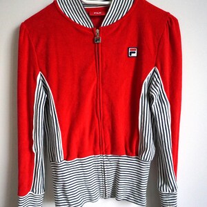 May include: A red zip-up jacket with white and black striped accents. The jacket features a white and black striped collar, cuffs, and lower hem. The Fila logo is visible on the chest. The jacket is hanging on a white hanger.