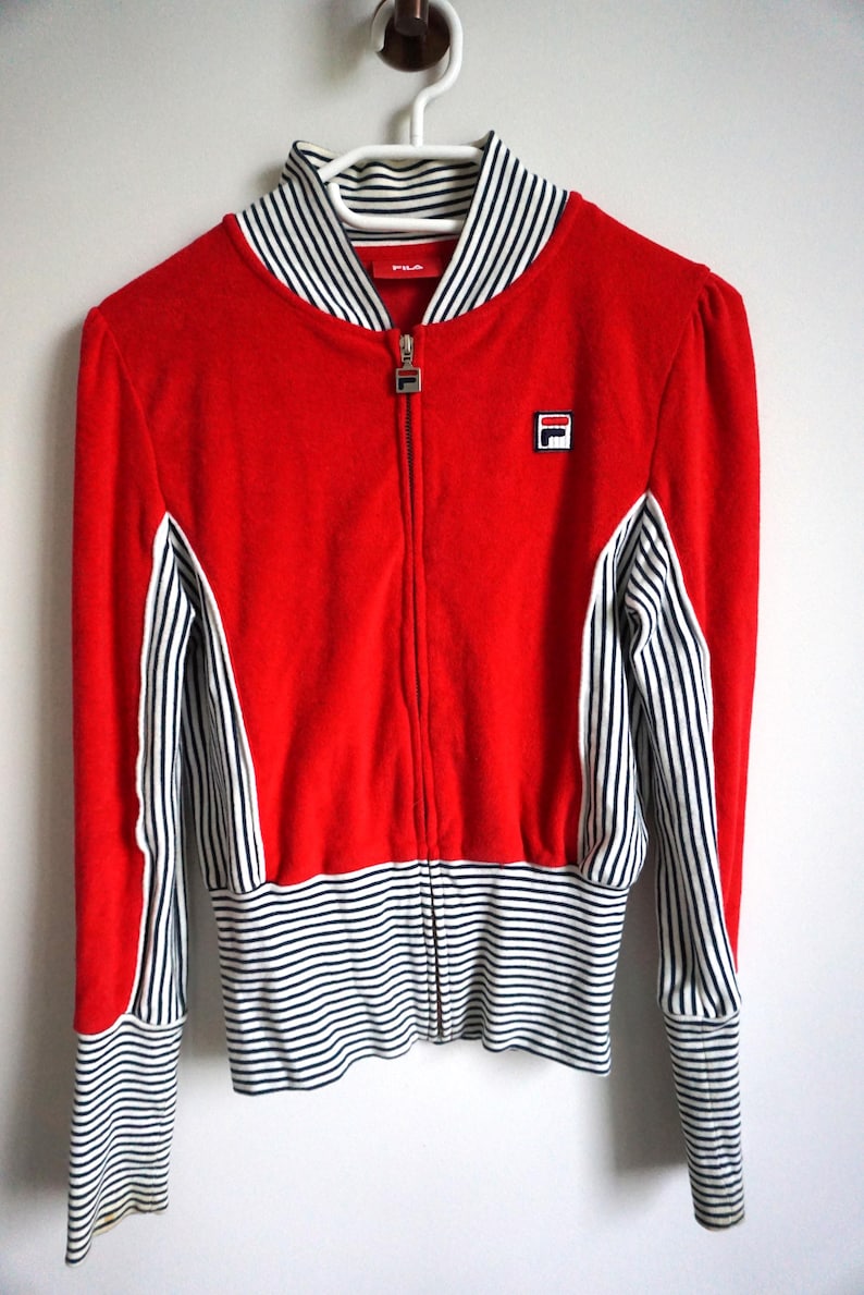 May include: A red zip-up jacket with white and black striped accents. The jacket features a white and black striped collar, cuffs, and lower hem. The Fila logo is visible on the chest. The jacket is hanging on a white hanger.