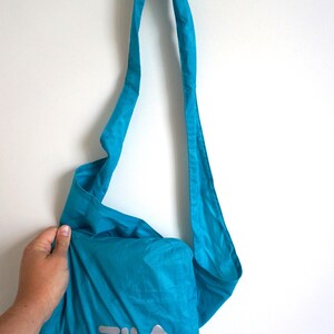 Pode incluir: Uma bolsa tiracolo turquesa brilhante feita de tecido leve e amassado. A bolsa possui uma al&ccedil;a de ombro longa e ajust&aacute;vel e a palavra FILA impressa em letras prateadas refletivas grandes na parte inferior frontal da bolsa.