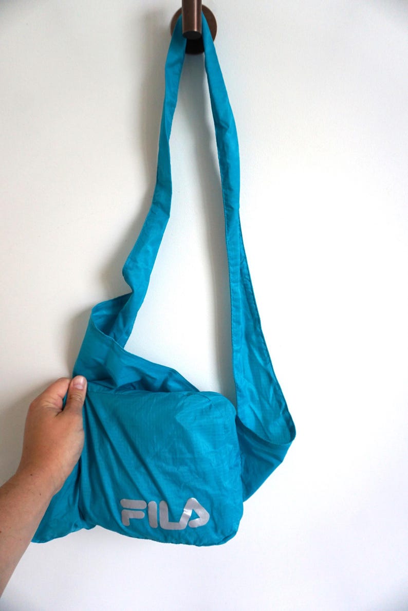 Pode incluir: Uma bolsa tiracolo turquesa brilhante feita de tecido leve e amassado. A bolsa possui uma al&ccedil;a de ombro longa e ajust&aacute;vel e a palavra FILA impressa em letras prateadas refletivas grandes na parte inferior frontal da bolsa.