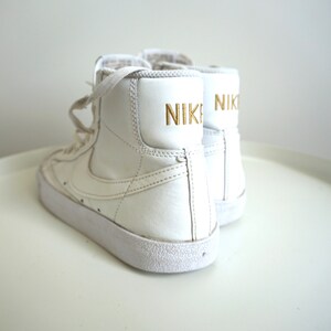 Op de afbeelding: Een paar witte high-top sneakers met het woord "NIKE" in gouden letters op de achterkant. De schoenen hebben witte veters en een witte zool. De sneakers zijn een klassieke atletische stijl.