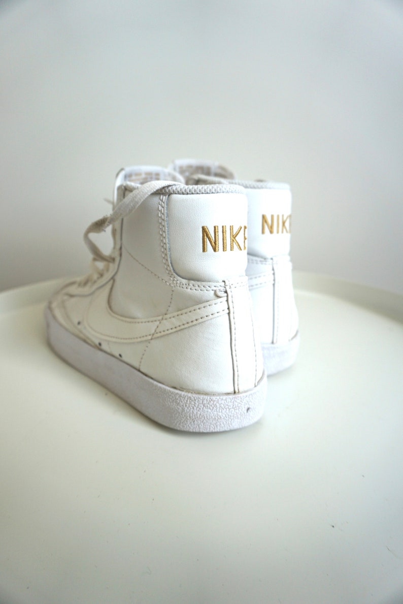 Op de afbeelding: Een paar witte high-top sneakers met het woord "NIKE" in gouden letters op de achterkant. De schoenen hebben witte veters en een witte zool. De sneakers zijn een klassieke atletische stijl.