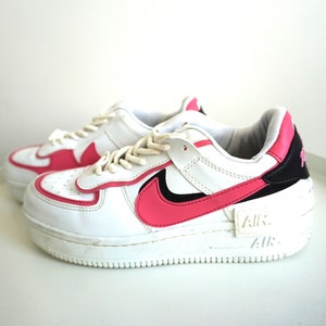 Puede incluir: Zapatillas Nike Air Force 1 blancas y rosas. Las zapatillas tienen una parte superior de cuero blanco con detalles en rosa en el logotipo de la marca, el tal&oacute;n y el ribete. La suela es blanca y la palabra "AIR" es visible en el lateral.