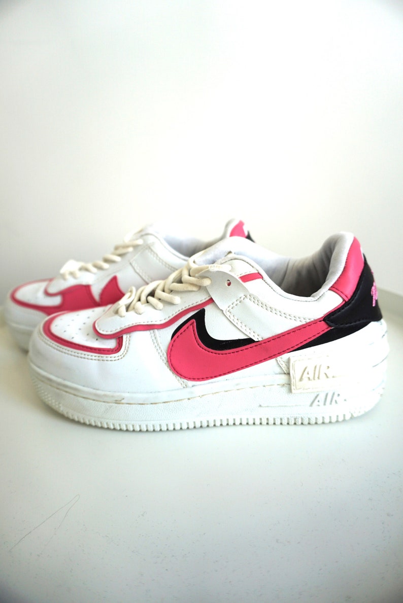 Puede incluir: Zapatillas Nike Air Force 1 blancas y rosas. Las zapatillas tienen una parte superior de cuero blanco con detalles en rosa en el logotipo de la marca, el tal&oacute;n y el ribete. La suela es blanca y la palabra "AIR" es visible en el lateral.