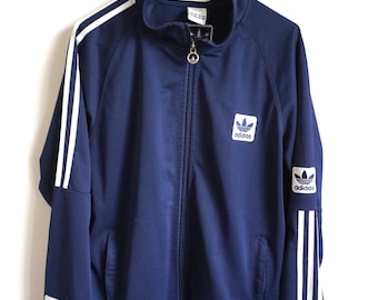 May include: Navy blue track jacket with white stripes on the sleeves and the Adidas logo on the chest and sleeve. The jacket has a full zipper and two front pockets. The jacket is hanging on a white hanger.