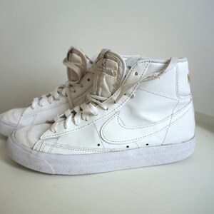 Op de afbeelding: Paar witte high-top sneakers met veters. De sneakers hebben een witte zool en een Nike swoosh-logo aan de zijkant. De tong en kraag zijn gewatteerd. Het Nike-logo is goudkleurig op de achterkant van de schoen.