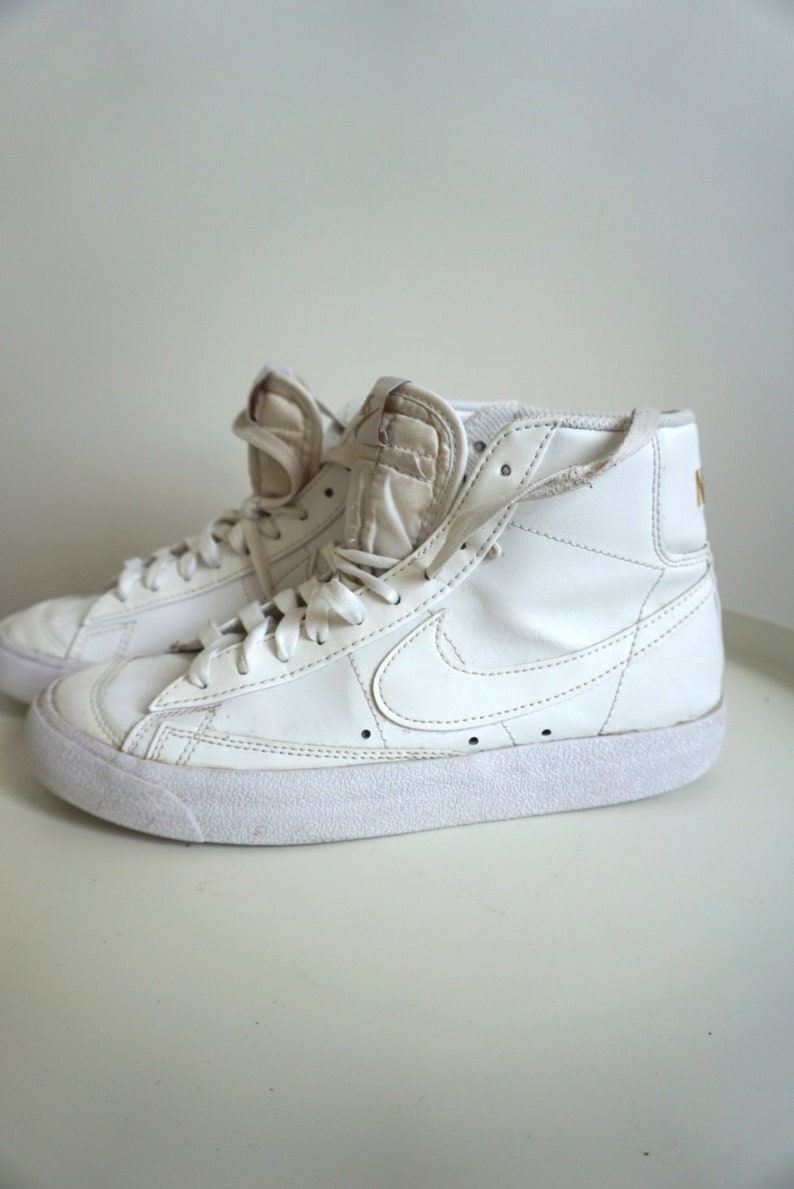 Op de afbeelding: Paar witte high-top sneakers met veters. De sneakers hebben een witte zool en een Nike swoosh-logo aan de zijkant. De tong en kraag zijn gewatteerd. Het Nike-logo is goudkleurig op de achterkant van de schoen.