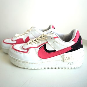 Puede incluir: Zapatillas blancas y rosas con suela gruesa. Los zapatos presentan un dise&ntilde;o de swoosh y detalles en rosa, con detalles en negro en el tal&oacute;n. La palabra "AIR" es visible en el lateral de la suela. Los cordones son blancos.