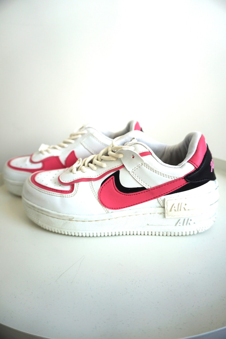 Puede incluir: Zapatillas blancas y rosas con suela gruesa. Los zapatos presentan un dise&ntilde;o de swoosh y detalles en rosa, con detalles en negro en el tal&oacute;n. La palabra "AIR" es visible en el lateral de la suela. Los cordones son blancos.