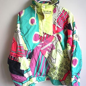 Puede incluir: Una chaqueta cortavientos de manga larga con un estampado gráfico abstracto multicolor en amarillo neón, verde azulado, magenta y negro. La chaqueta incluye un cuello alto con cordón, un bolsillo frontal con cremallera y botones a presión.