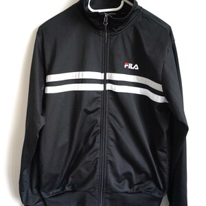 May include: A black track jacket with a high collar and a full-length front zipper. The jacket features two horizontal white stripes across the chest and the FILA logo embroidered in white and red on the upper left side of the chest area.