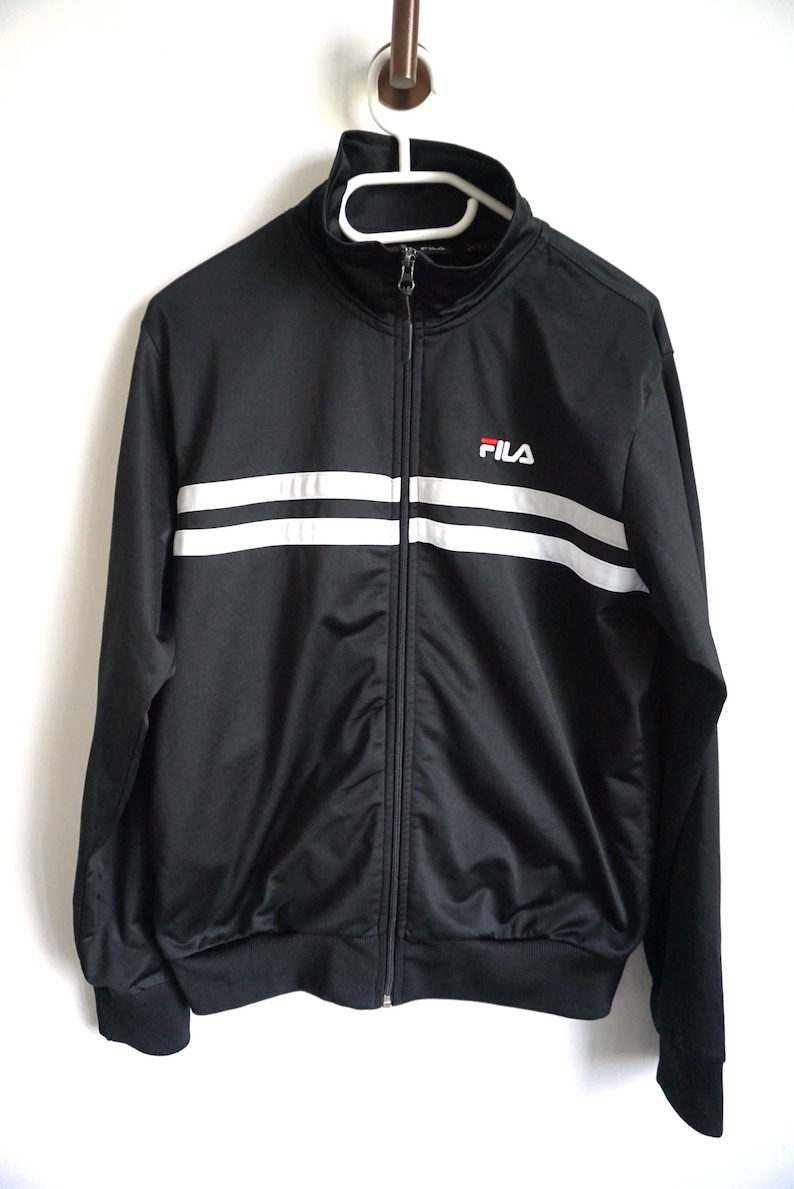 May include: A black track jacket with a high collar and a full-length front zipper. The jacket features two horizontal white stripes across the chest and the FILA logo embroidered in white and red on the upper left side of the chest area.