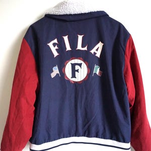 K&ouml;nnte beinhalten: Eine marineblaue und rote Fila-Jacke mit wei&szlig;em Sherpa-Kragen. Auf der R&uuml;ckseite steht in Wei&szlig; das Wort "FILA", zusammen mit einem gro&szlig;en "F"-Logo und Flaggen. Die &Auml;rmel sind rot, und die Jacke hat einen wei&szlig;-blauen Streifen am Saum.