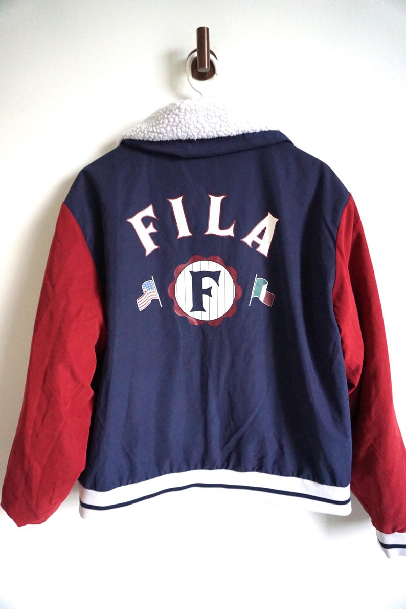 K&ouml;nnte beinhalten: Eine marineblaue und rote Fila-Jacke mit wei&szlig;em Sherpa-Kragen. Auf der R&uuml;ckseite steht in Wei&szlig; das Wort "FILA", zusammen mit einem gro&szlig;en "F"-Logo und Flaggen. Die &Auml;rmel sind rot, und die Jacke hat einen wei&szlig;-blauen Streifen am Saum.