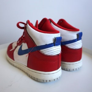 Puede incluir: Zapatillas altas rojas, blancas y azules. Las zapatillas tienen una parte superior de cuero rojo con detalles blancos y azules, una entresuela blanca y un logotipo azul de la marca. Los cordones son rojos.