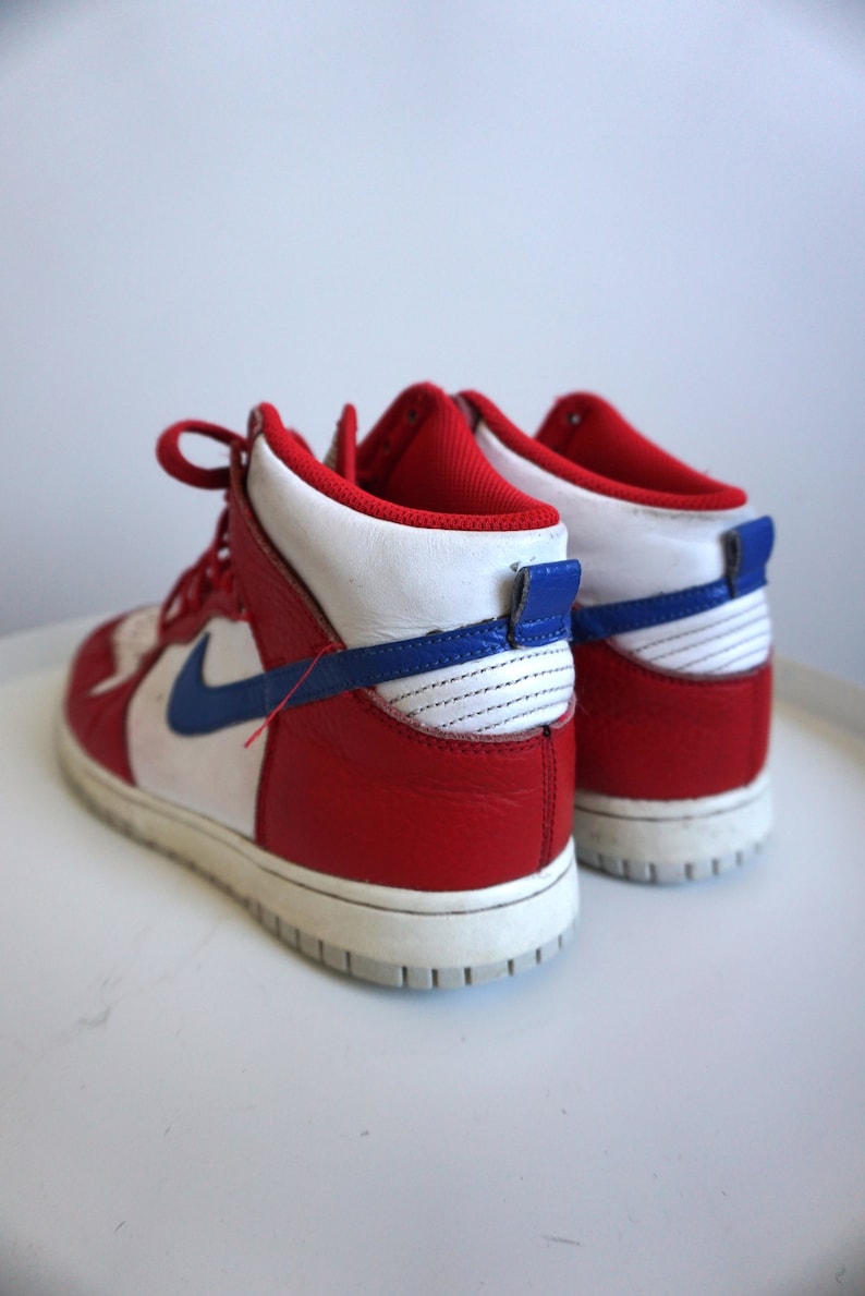 Puede incluir: Zapatillas altas rojas, blancas y azules. Las zapatillas tienen una parte superior de cuero rojo con detalles blancos y azules, una entresuela blanca y un logotipo azul de la marca. Los cordones son rojos.