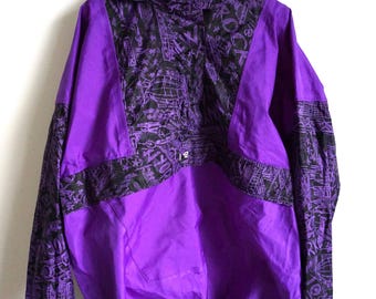 May include: A purple hooded jacket with black patterned accents. The jacket features a drawstring at the hem and black cuffs. The black pattern includes various shapes and designs. The jacket is hanging on a white hanger.