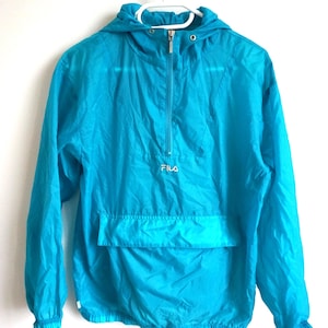Pode incluir: Um blus&atilde;o corta-vento tipo pullover em turquesa brilhante com capuz, mangas compridas com punhos el&aacute;sticos e fecho de meio z&iacute;per frontal. O log&oacute;tipo FILA est&aacute; bordado a branco no centro do peito, acima de um grande bolso frontal.