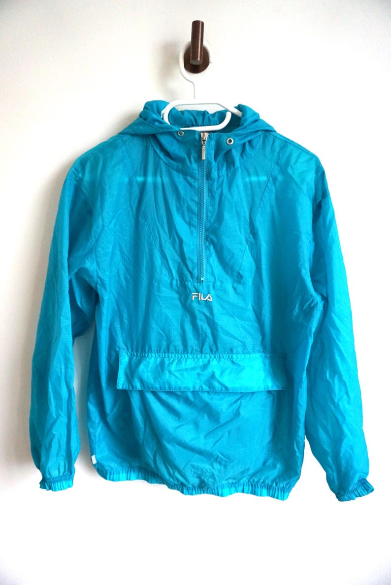 Pode incluir: Um blus&atilde;o corta-vento tipo pullover em turquesa brilhante com capuz, mangas compridas com punhos el&aacute;sticos e fecho de meio z&iacute;per frontal. O log&oacute;tipo FILA est&aacute; bordado a branco no centro do peito, acima de um grande bolso frontal.